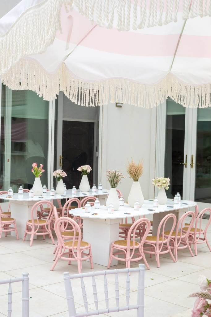 Pink rattan kids' chairs at a long white table under a fringed umbrella on a poolside patio